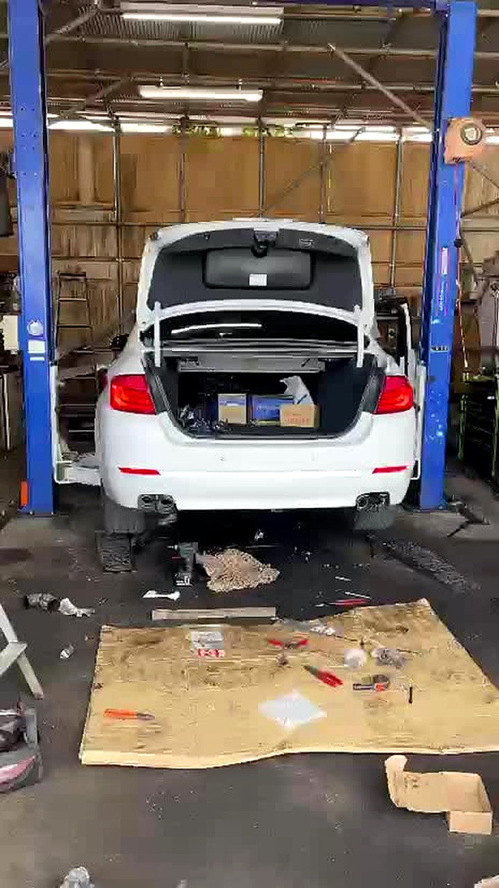 White BMW 523i F10 sedan on a blue two-post lift in a workshop, boot open, red-jacketed technician working at the right rear wheel, valved SUS304 catback fitted, no number plate visible