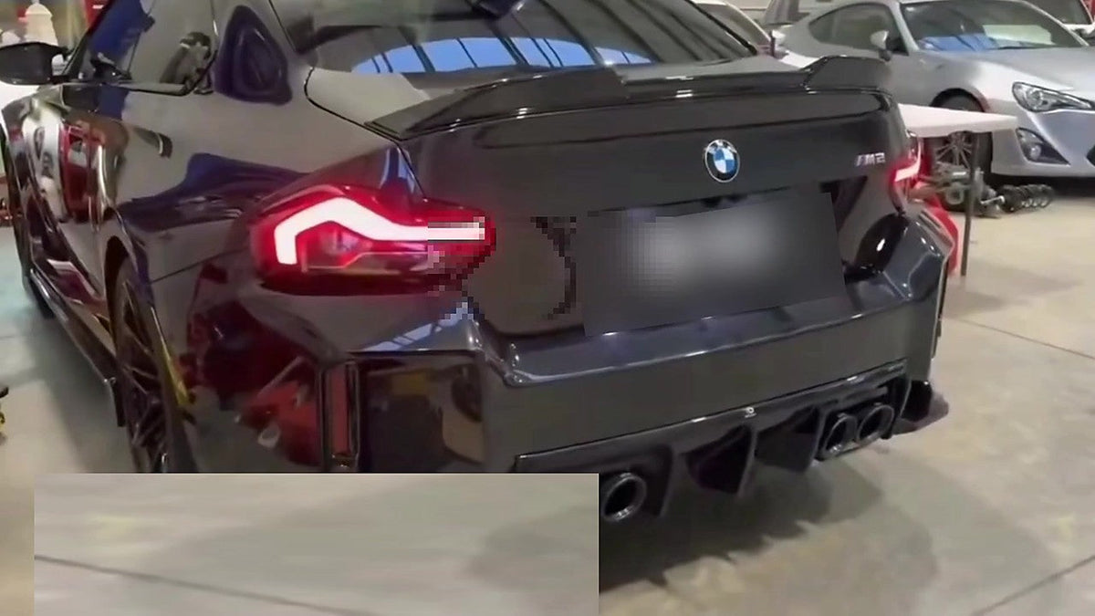 Black BMW M2 G87 rear three-quarter view in a workshop, carbon ducktail spoiler, lit OLED taillight, four black exhaust tips in the diffuser, number plate pixelated