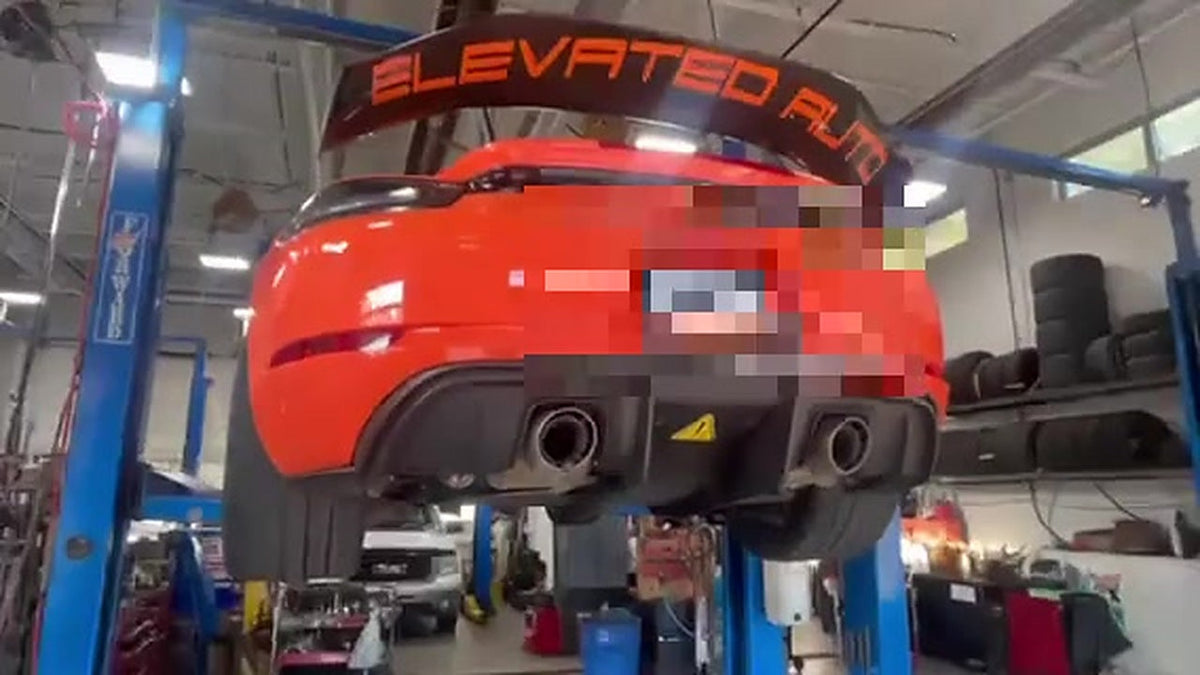 Orange Porsche 718 GT4 RS raised on a workshop lift, rear bumper and GT4 RS wing in view, number plate pixelated for privacy