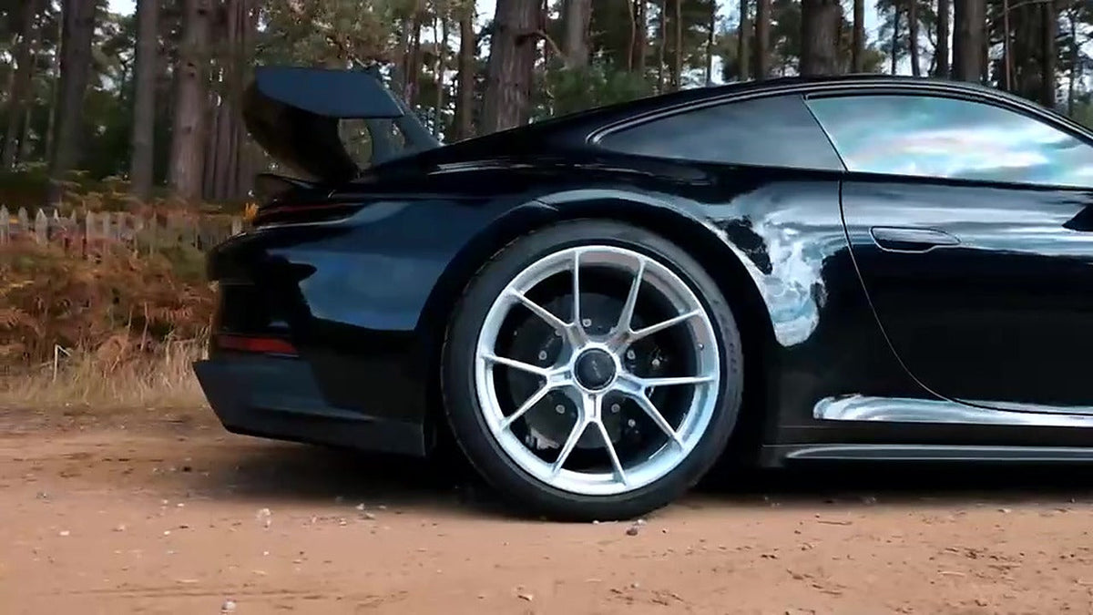 Black Porsche 911 992 GT3 side profile on a forest lane, silver centre-lock wheels and swan-neck rear wing visible, number plate not in frame